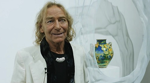 Artist David Begbie is interviewed during a sculpture exhibition. He is standing in front of a wire-mesh sculpture and an antique colourful vase.